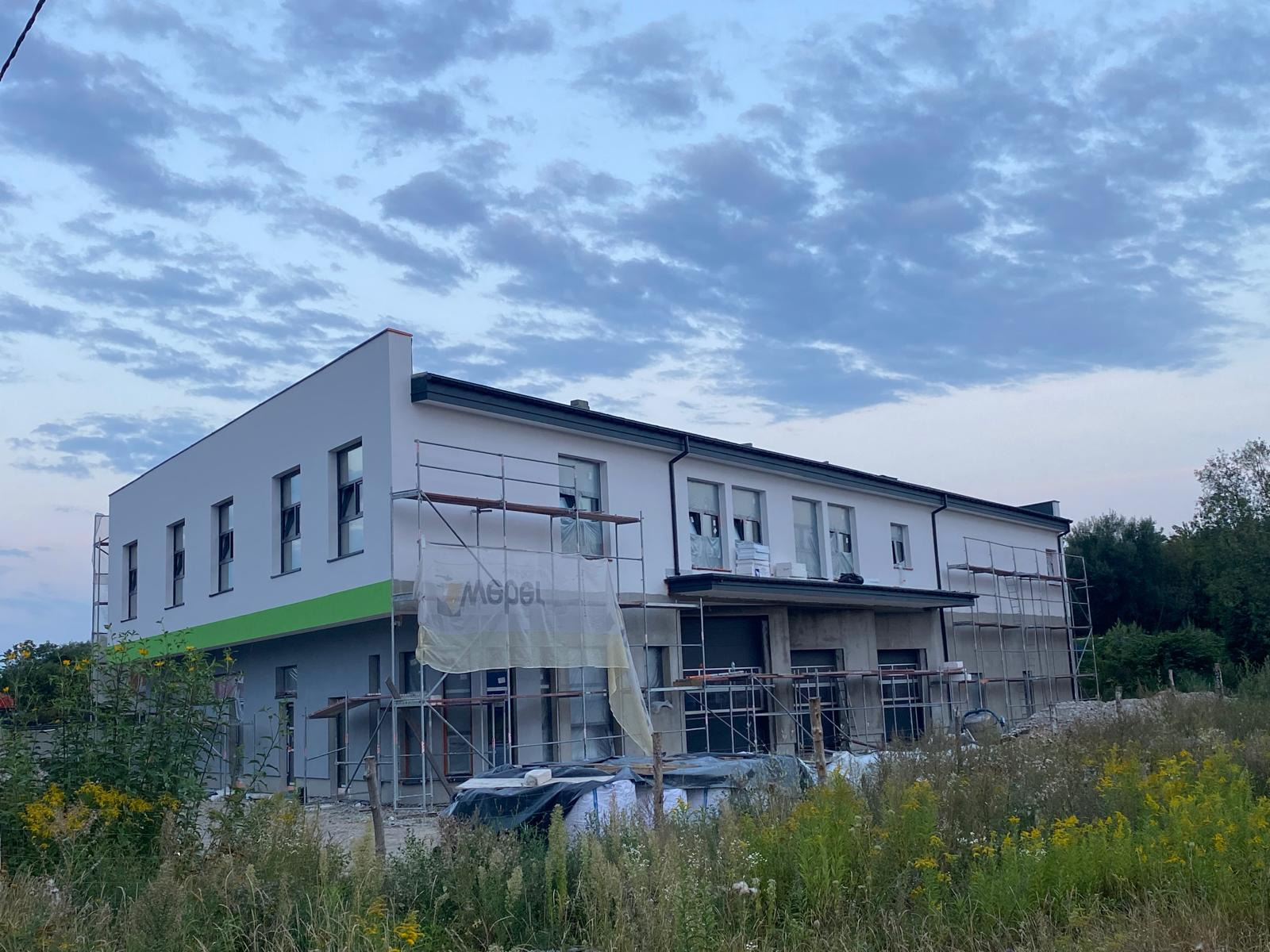 Dwukondygnacyjny budynek w trakcie ocieplania elewacji, widoczne rusztowania i zabezpieczenia. Częściowo otynkowana fasada w jasnych barwach, zieleń w tle.