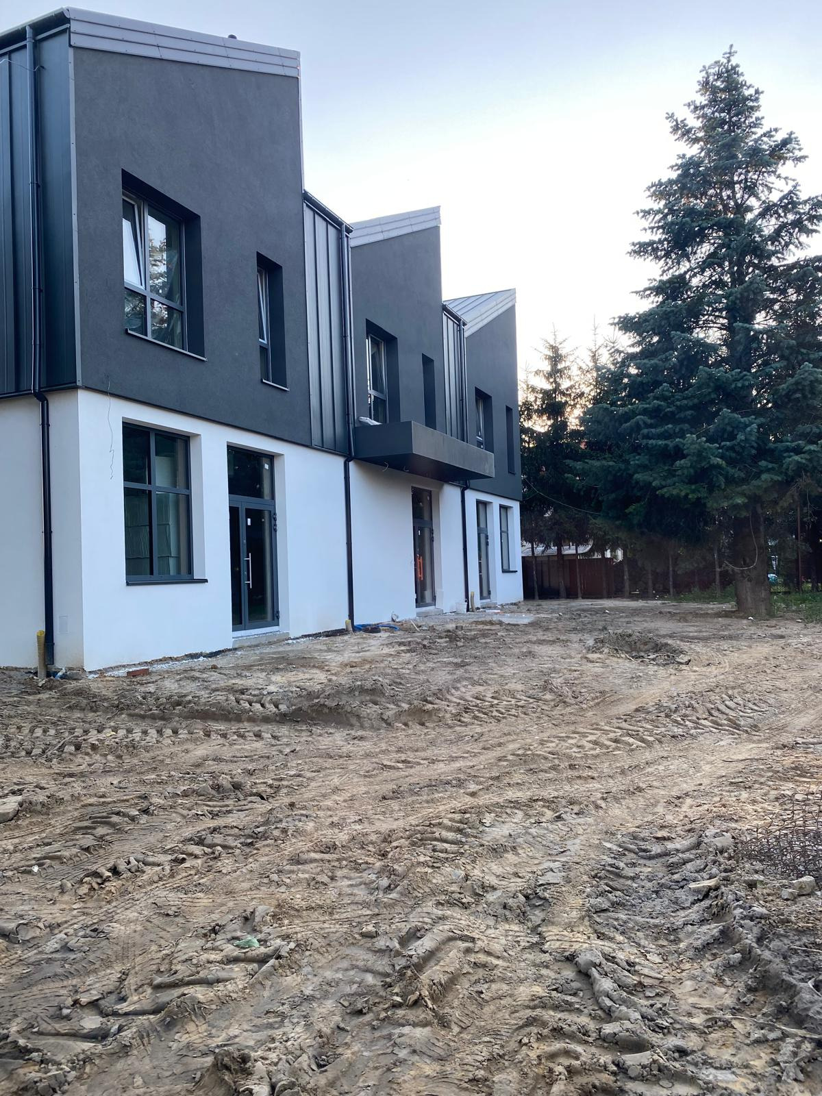 Nowoczesny budynek z szarą elewacją i białymi ścianami parteru, duże okna, teren wokół w trakcie prac ziemnych, wysokie drzewa w tle.