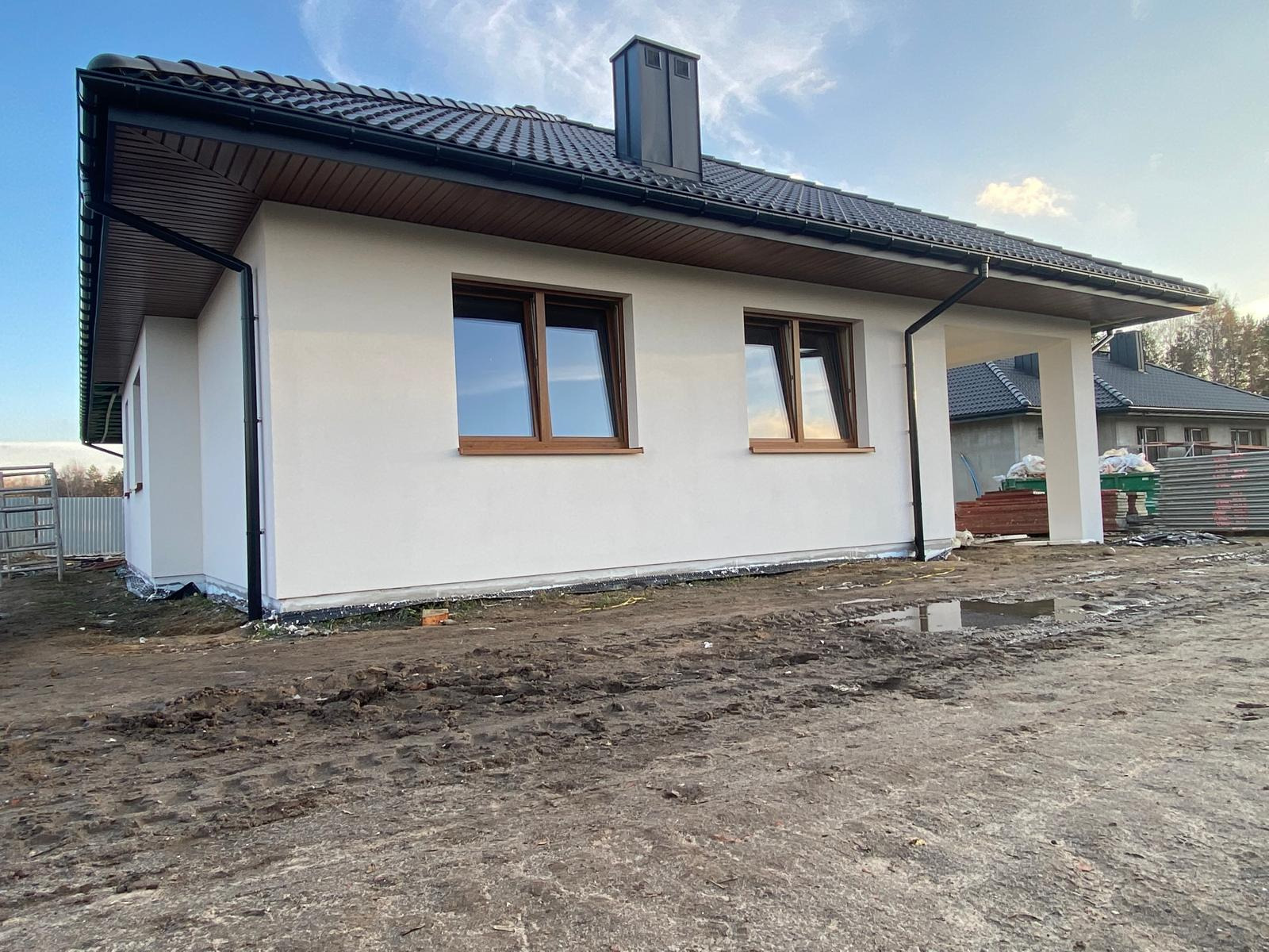 Nowoczesny dom z białą elewacją, brązowymi oknami i ciemną dachówką. Widoczne ślady prac ziemnych wokół budynku. Minimalistyczny design z wyraźnymi liniami.