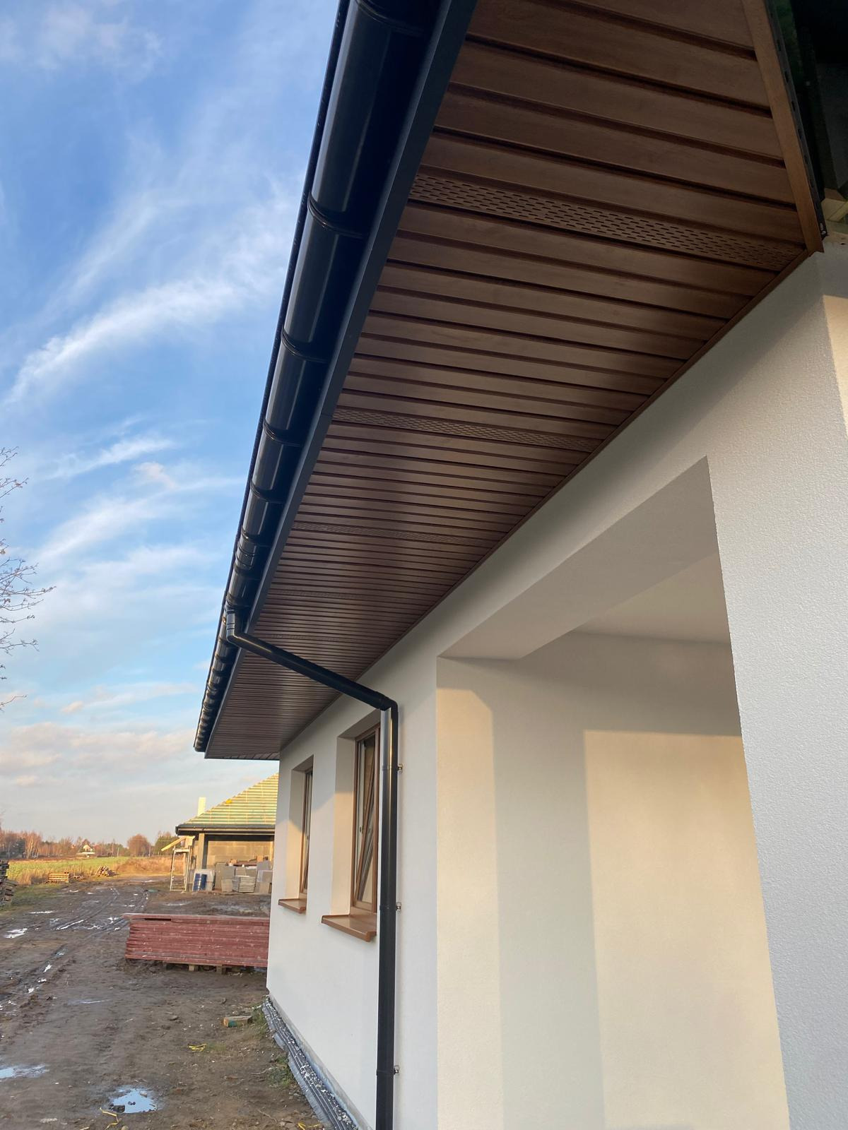 Elewacja domu jednorodzinnego w jasnym kolorze, z drewnianą podbitką i orynnowaniem. Widok z dołu, na tle błękitnego nieba.