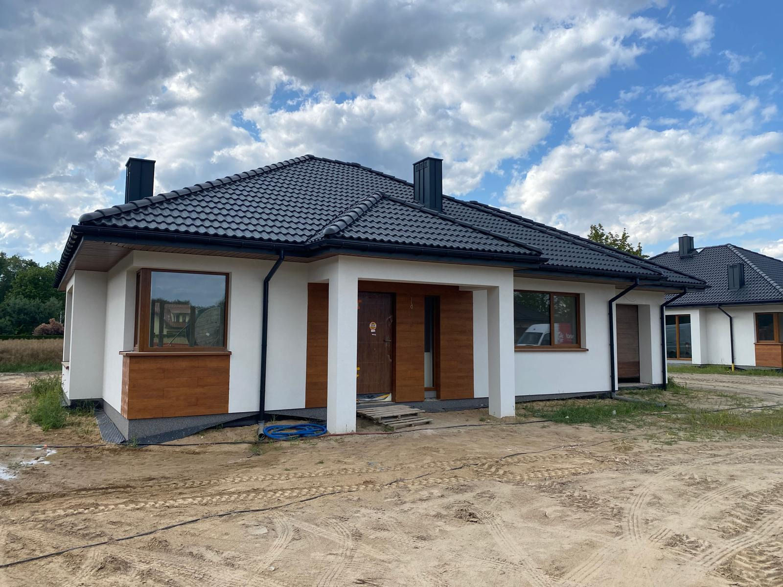 Nowoczesny dom jednorodzinny z białą elewacją i drewnianymi akcentami w Warszawie. Czarny dach z dachówki ceramicznej, widoczne rynny i komin.