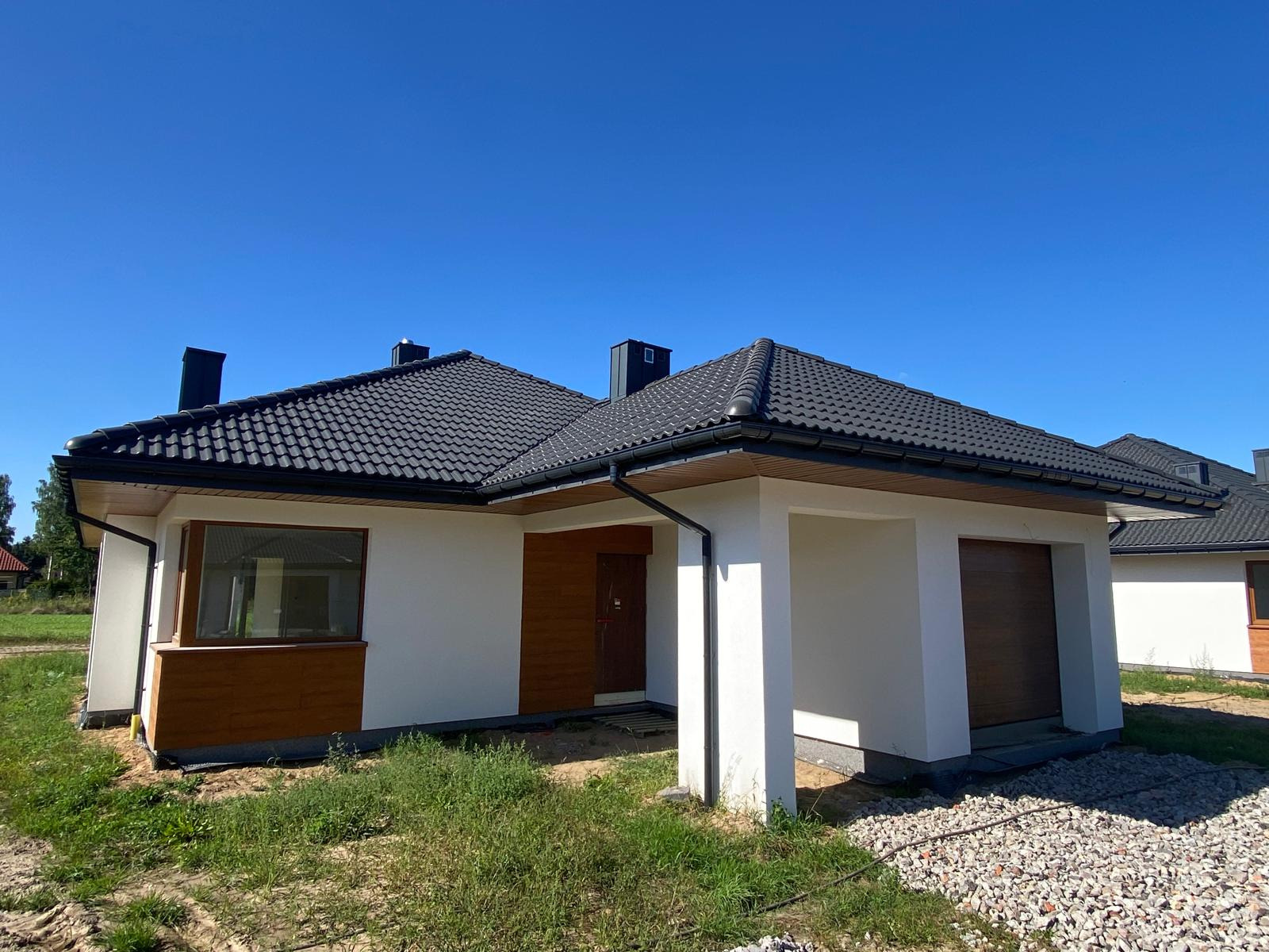 Nowy dom jednorodzinny z ciemną dachówką i białymi ścianami, z drewnianymi akcentami, w słoneczny dzień. Widoczny fragment garażu i starannie wykonana elewacja.