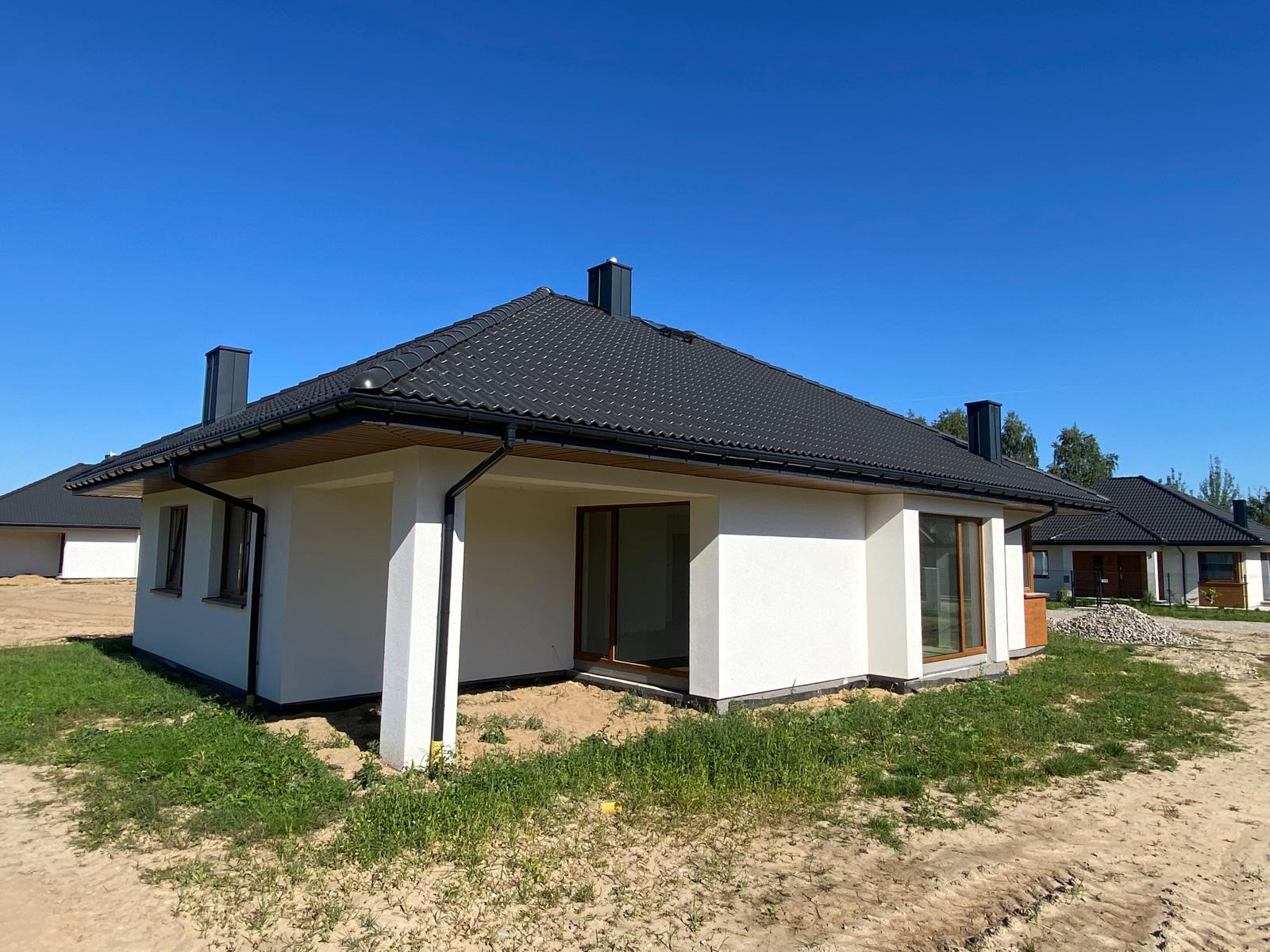 Nowoczesny dom jednorodzinny z białą elewacją i ciemną dachówką, widok z zewnątrz w słoneczny dzień. Dom w stanie deweloperskim, z widocznymi elementami wykończeniowymi.