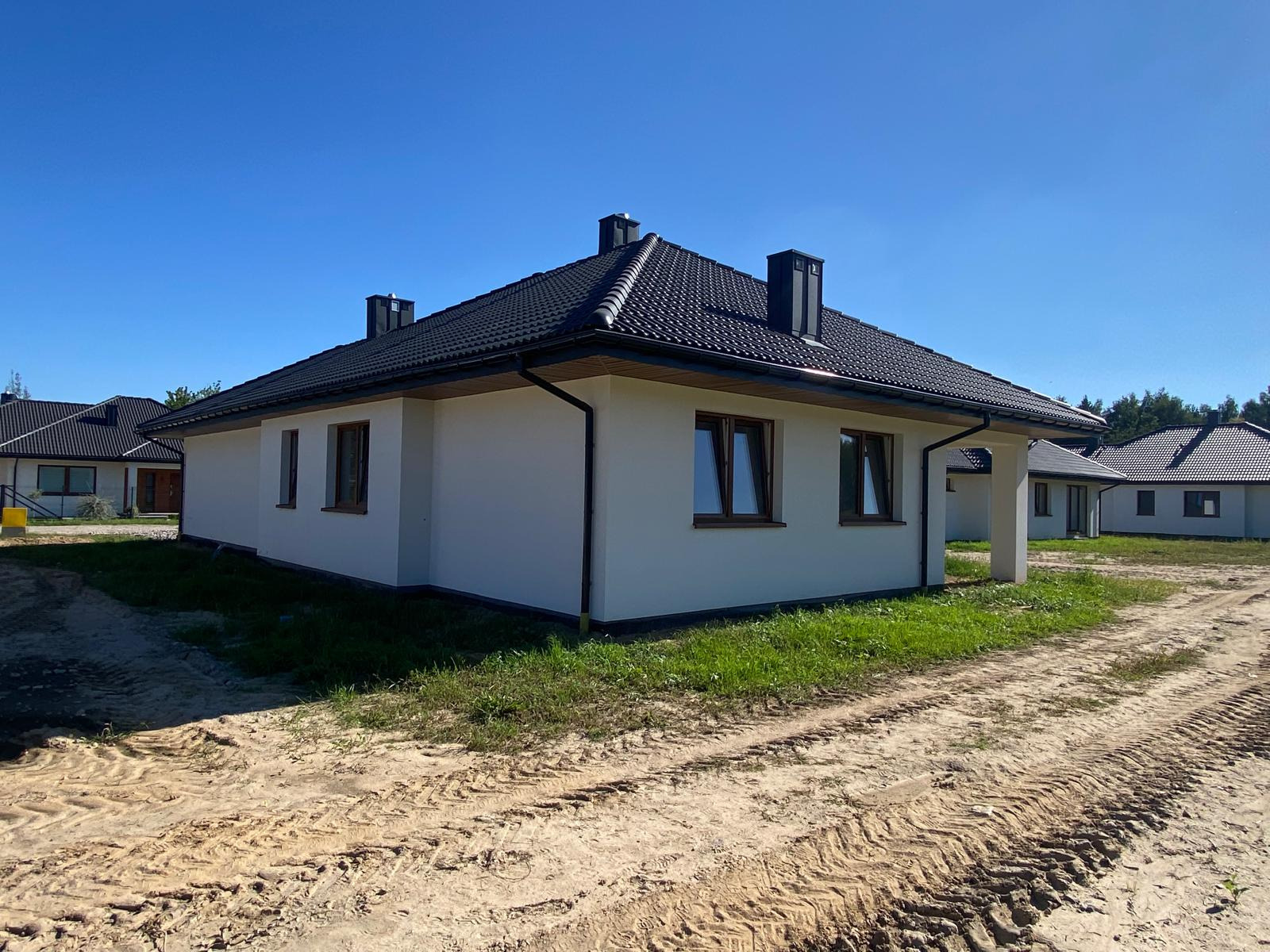 Nowoczesny, parterowy dom z białą elewacją i czarnym dachem z dachówki, widok z zewnątrz na tle błękitnego nieba i innych domów w oddali. Ślady opon na piaszczystym podłożu.