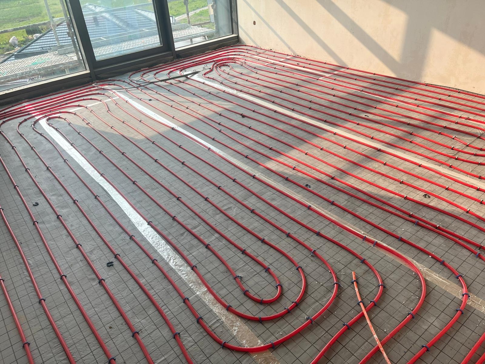Czerwone rury ogrzewania podłogowego ułożone równolegle na szarej siatce w jasnym pomieszczeniu z dużym oknem. Widoczne mocowania rur.