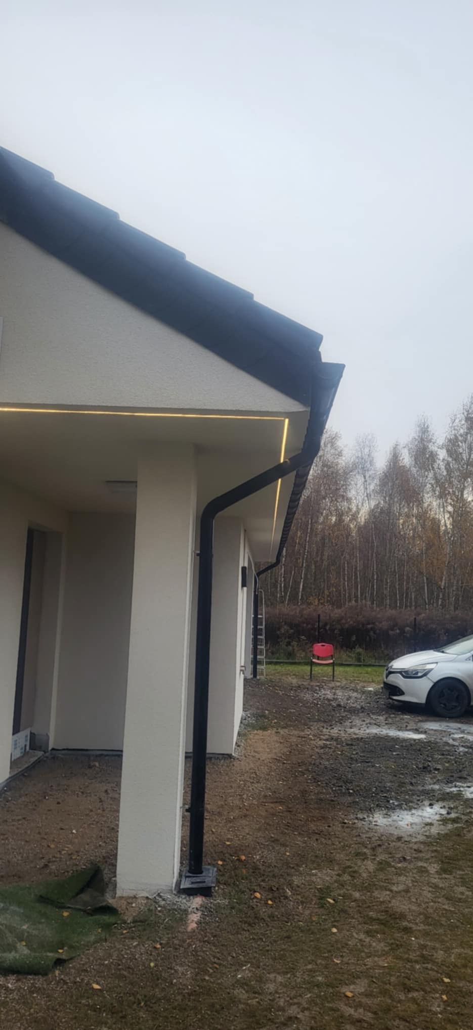 Elewacja domu z oświetleniem LED pod okapem i czarną rynną. Widoczny fragment samochodu i drzewa w tle. Ujęcie z dołu, podkreślające linię światła.