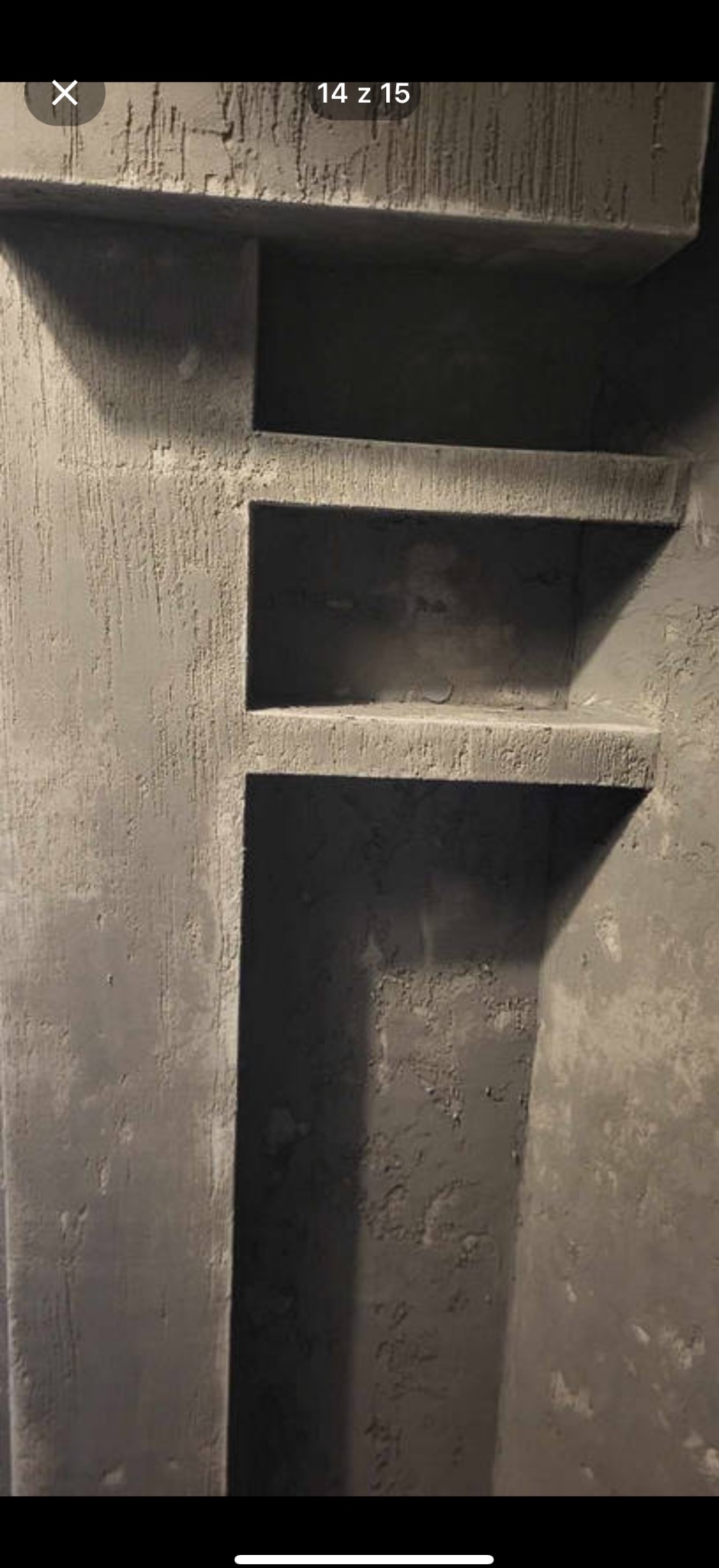 Surowa ściana z wnękami o szorstkiej fakturze, w odcieniach szarości, z widocznymi śladami prac wykończeniowych. Minimalistyczny design i pionowy układ wnęk.