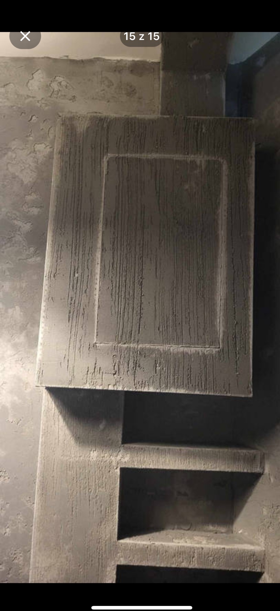 Wykończona, szara ściana z dekoracyjną strukturą, imitującą deszcz. Widoczne półki w ścianie i fragment szafki. Wyjątkowa faktura i surowy styl.