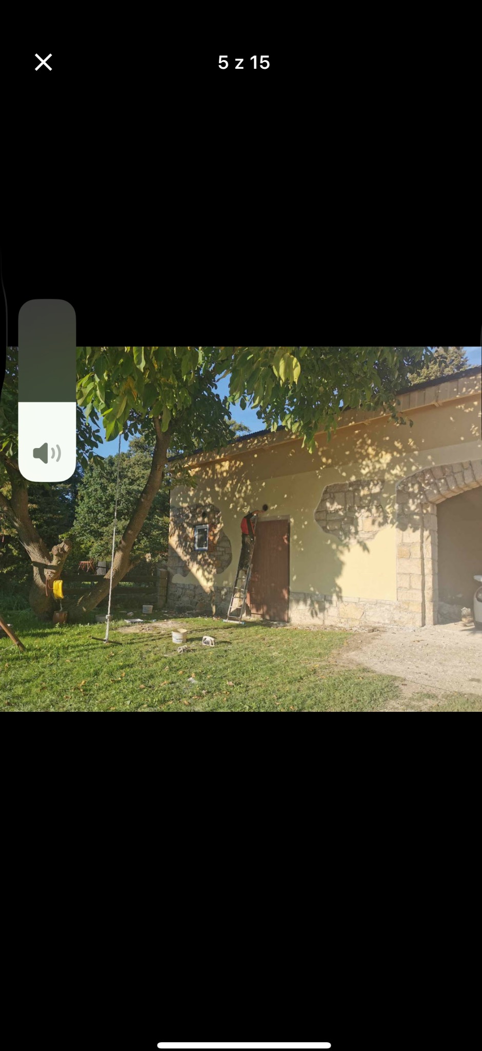 Malowanie elewacji budynku z elementami kamiennymi. Pracownik na drabinie, widoczne puszki z farbą, trawa, drzewo, słoneczny dzień.