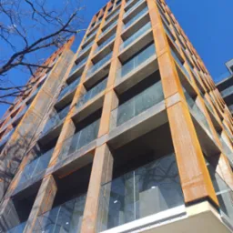 Wysoki budynek z cortenową elewacją i szklanymi balkonami, widziany z dołu pod ostrym kątem. Błękitne niebo i gałęzie drzew na pierwszym planie.