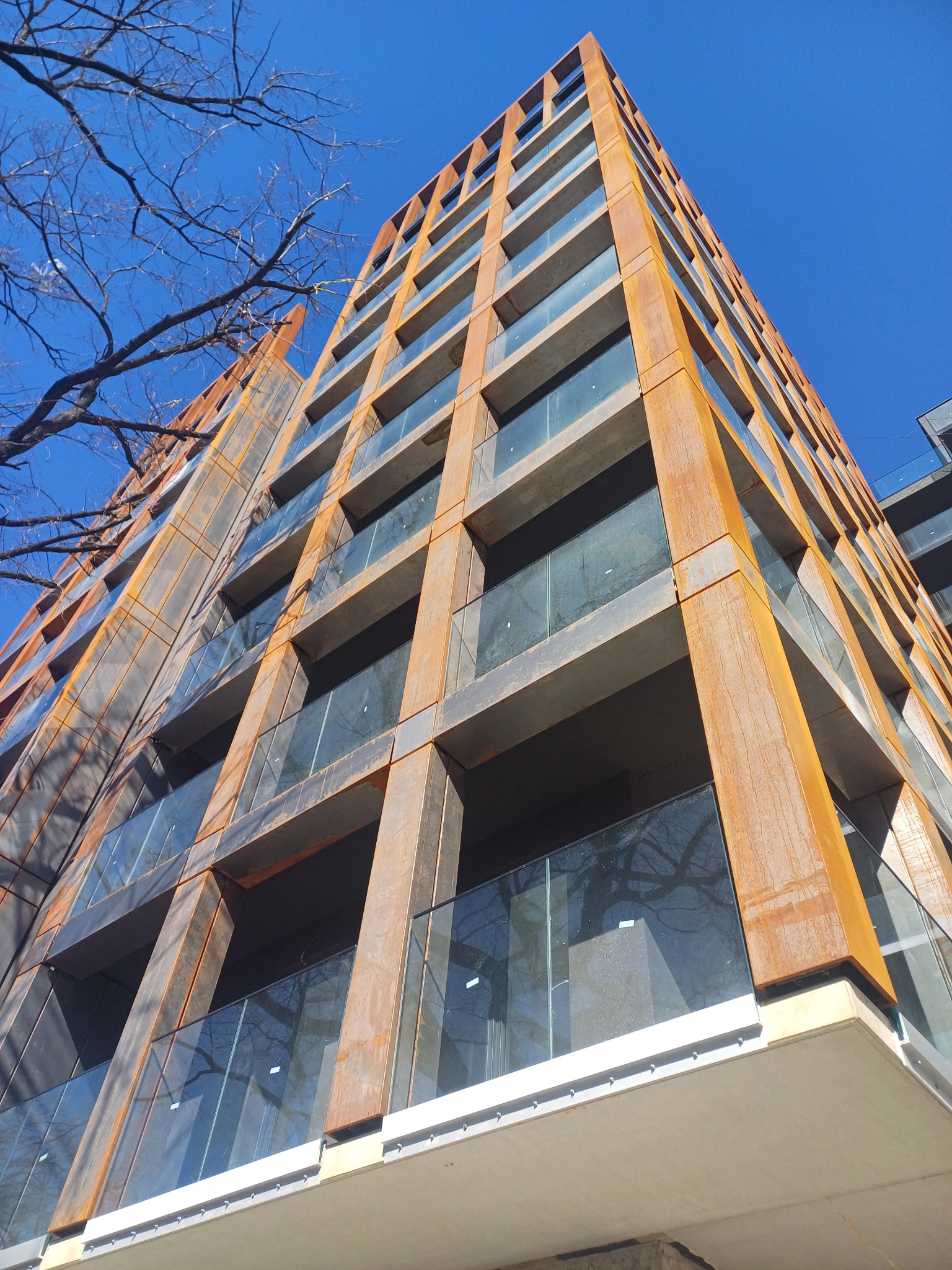 Wysoki budynek z cortenową elewacją i szklanymi balkonami, widziany z dołu pod ostrym kątem. Błękitne niebo i gałęzie drzew na pierwszym planie.