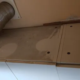 Brudna podszafkowa płyta z plamami, rura wentylacyjna i białe szafki. Widoczne ślady po czyszczeniu, kurz i pozostałości po remoncie. Perspektywa od dołu.