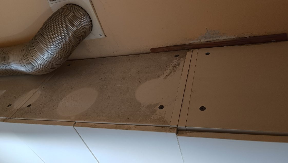 Brudna podszafkowa płyta z plamami, rura wentylacyjna i białe szafki. Widoczne ślady po czyszczeniu, kurz i pozostałości po remoncie. Perspektywa od dołu.