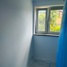 Usługi Remontowo-budowlane Grzegorz Fronia - Pomieszczenie po malowaniu na jasny odcień niebieskiego, z oknem i zabezpieczoną folią częścią podłogi. Widoczne świeżo pomalowane ściany i listwy przypodłogowe.