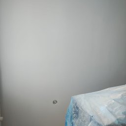 Usługi Remontowo-budowlane Grzegorz Fronia - Świeżo pomalowana, jasnoszara ściana z gniazdkiem elektrycznym i zabezpieczonym meblem folią ochronną. Widok z dołu, podkreślający gładkość wykończenia.