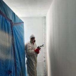 Usługi Remontowo-budowlane Grzegorz Fronia - Pracownik w kombinezonie ochronnym maluje ścianę na biało za pomocą pistoletu natryskowego. Widoczna niebieska folia ochronna i rury przy ścianie.