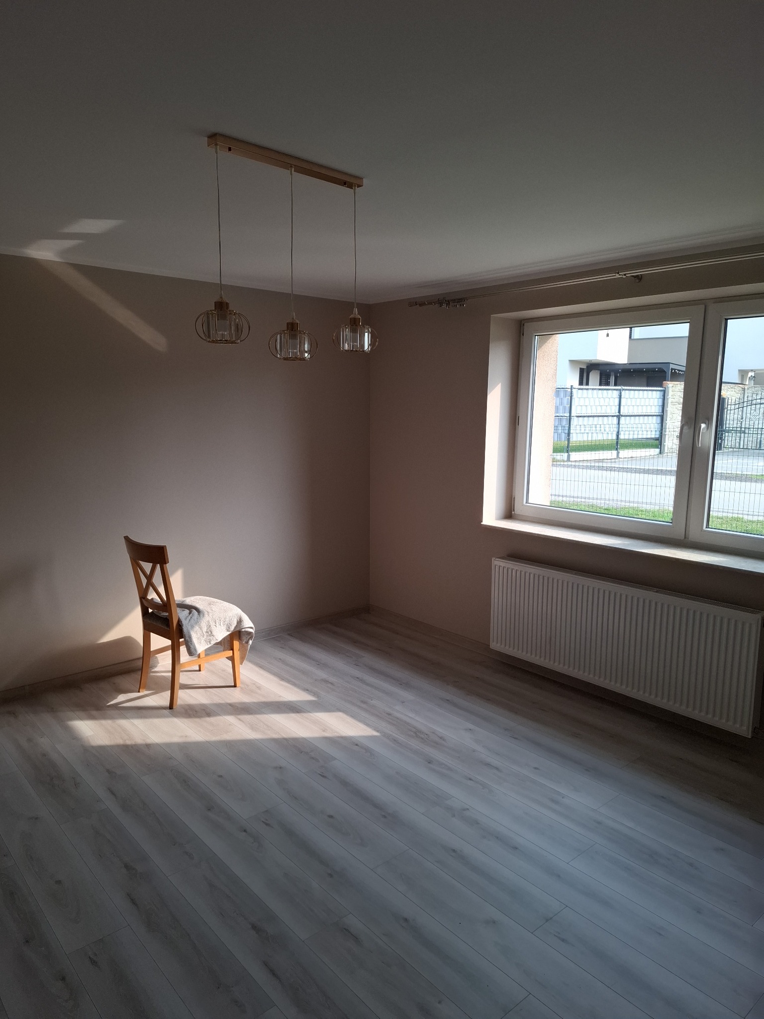Jasne, wykończone wnętrze pokoju z panelami podłogowymi, beżowymi ścianami, grzejnikiem pod oknem i krzesłem pod wiszącą lampą. Słoneczny blask na podłodze.