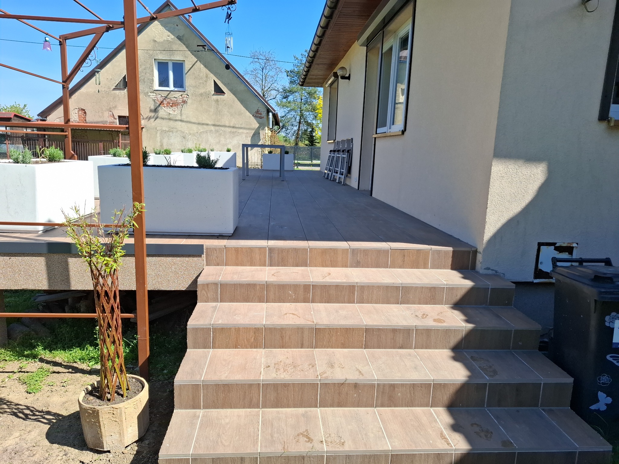 Zewnętrzne schody wyłożone brązowymi płytkami imitującymi drewno, prowadzące na taras z donicami i metalową pergolą. W tle budynek mieszkalny w słoneczny dzień.