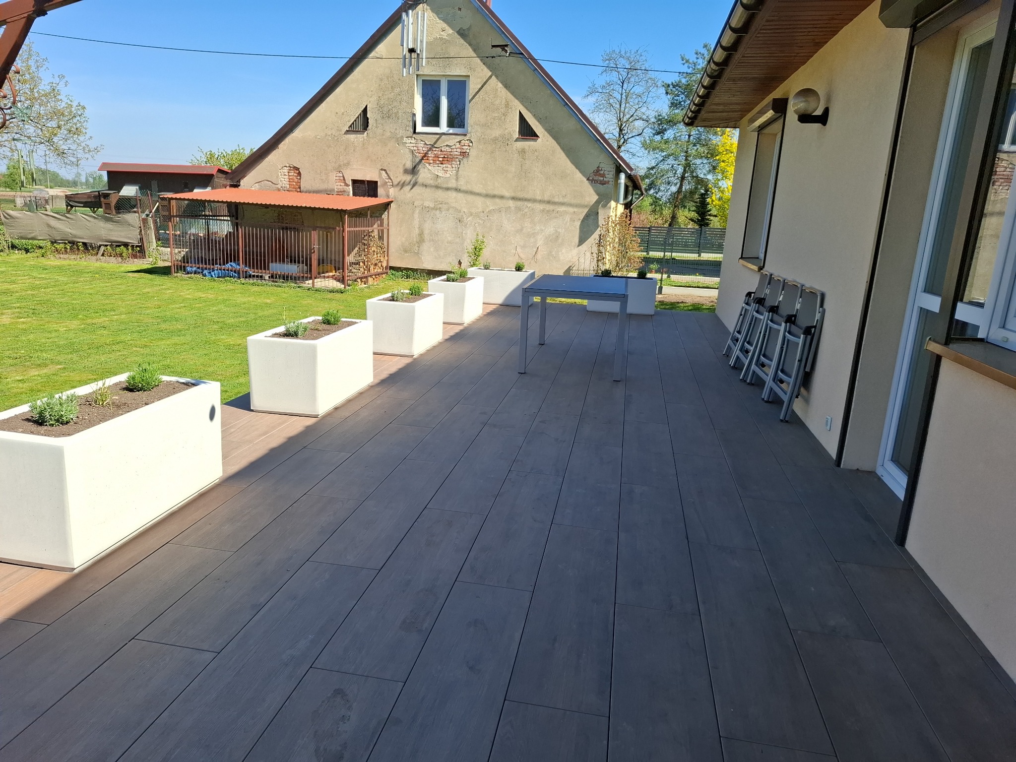 Nowoczesny taras z szarych desek kompozytowych, białych donic z zielenią i metalowym stolikiem. W tle dom z jasną elewacją i zielony trawnik.