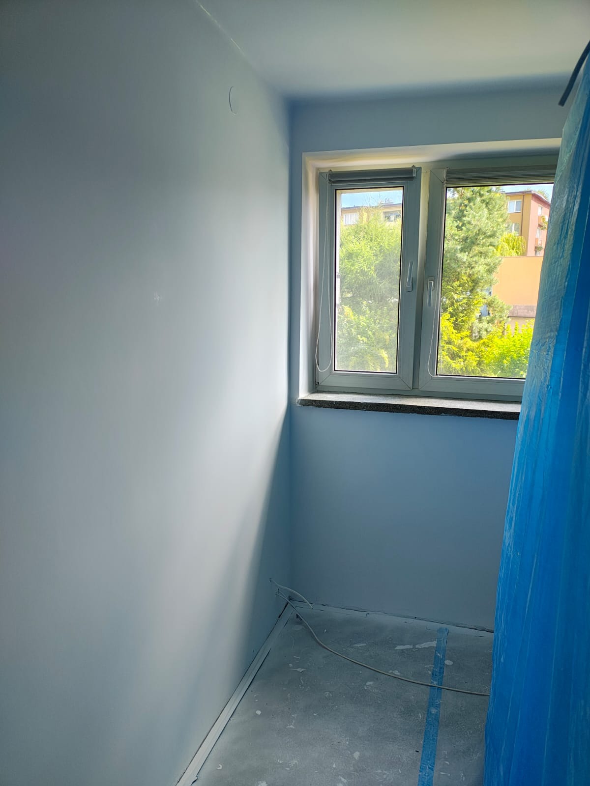 Pomieszczenie po malowaniu na jasny odcień niebieskiego, z oknem i zabezpieczoną folią częścią podłogi. Widoczne świeżo pomalowane ściany i listwy przypodłogowe.