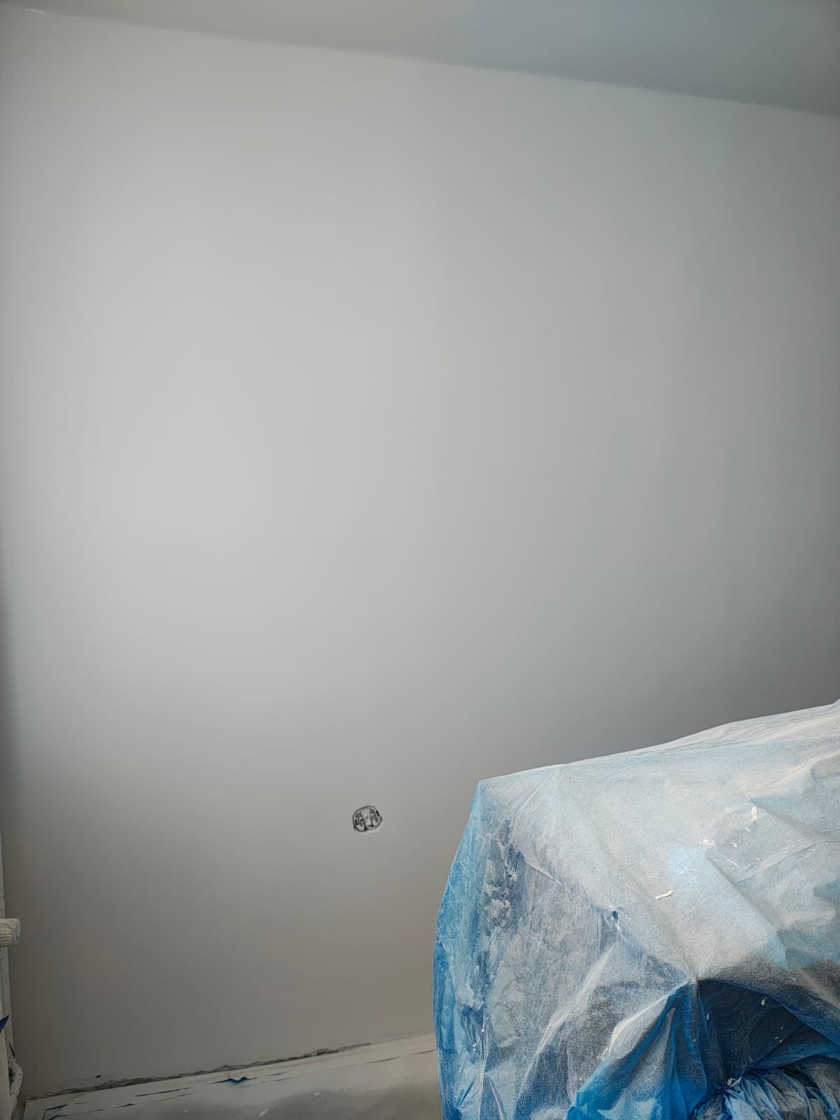 Świeżo pomalowana, jasnoszara ściana z gniazdkiem elektrycznym i zabezpieczonym meblem folią ochronną. Widok z dołu, podkreślający gładkość wykończenia.