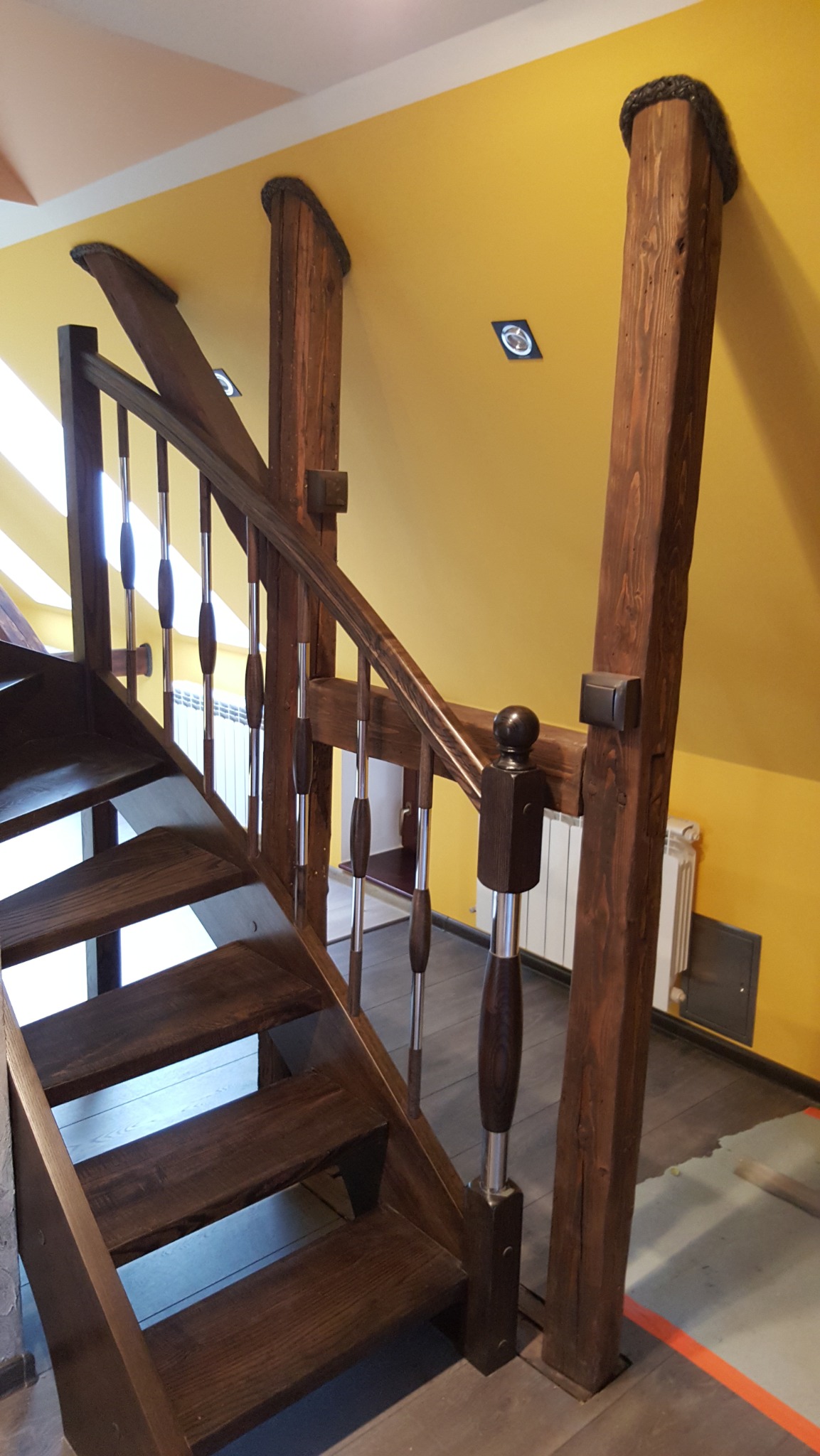 Drewniane schody z balustradą i ozdobnymi słupkami na tle żółtej ściany w skosie. Widoczne fragmenty podłogi i grzejnika.
