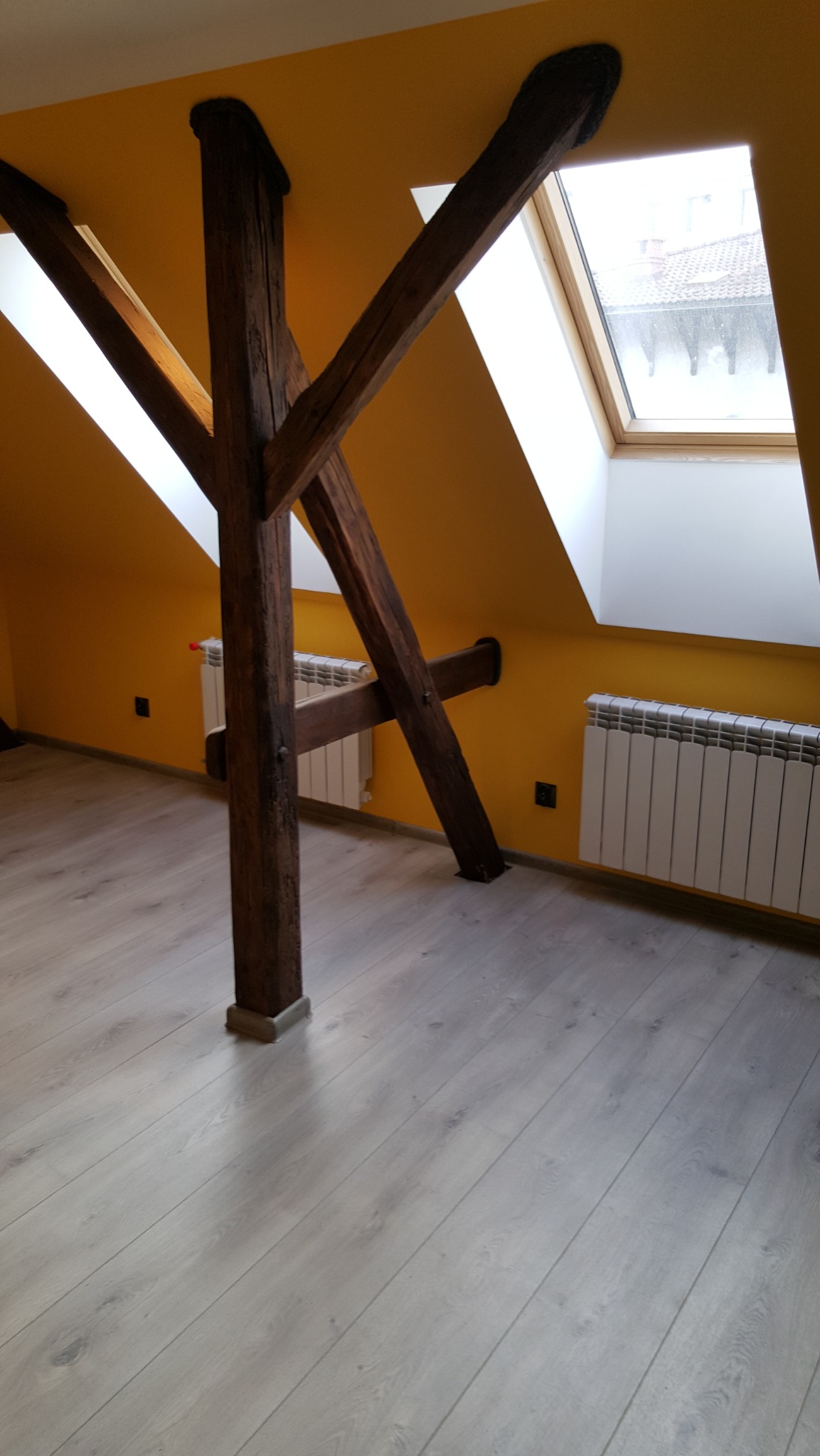 Żółte poddasze z drewnianą konstrukcją wspornikową, oknem dachowym i grzejnikami. Jasna podłoga z paneli. Charakterystyczny element architektoniczny.
