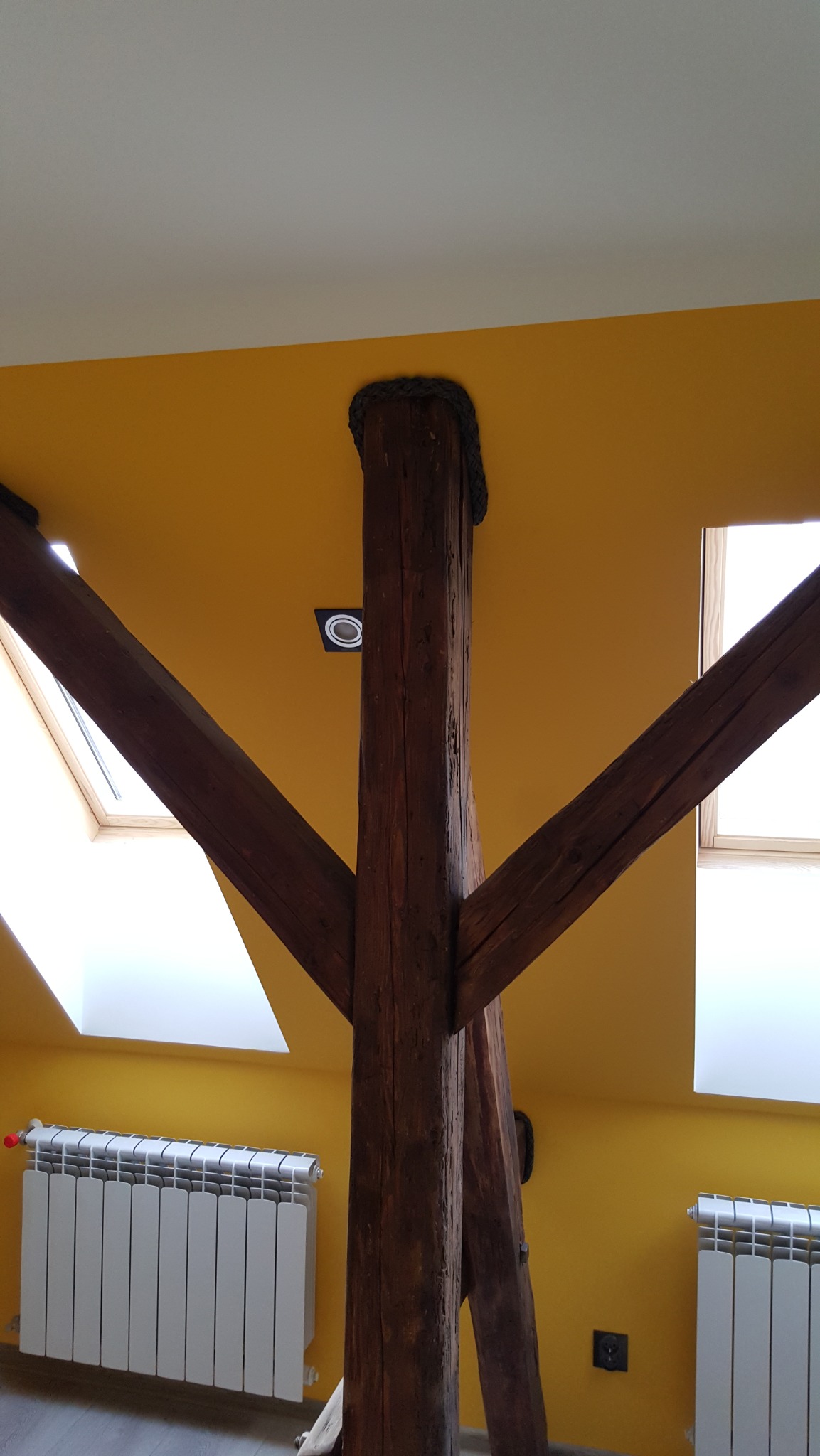 Żółte ściany poddasza z drewnianą kolumną i belkami, widoczne okna dachowe i grzejniki. Charakterystyczne wykończenie kolumny sznurem.