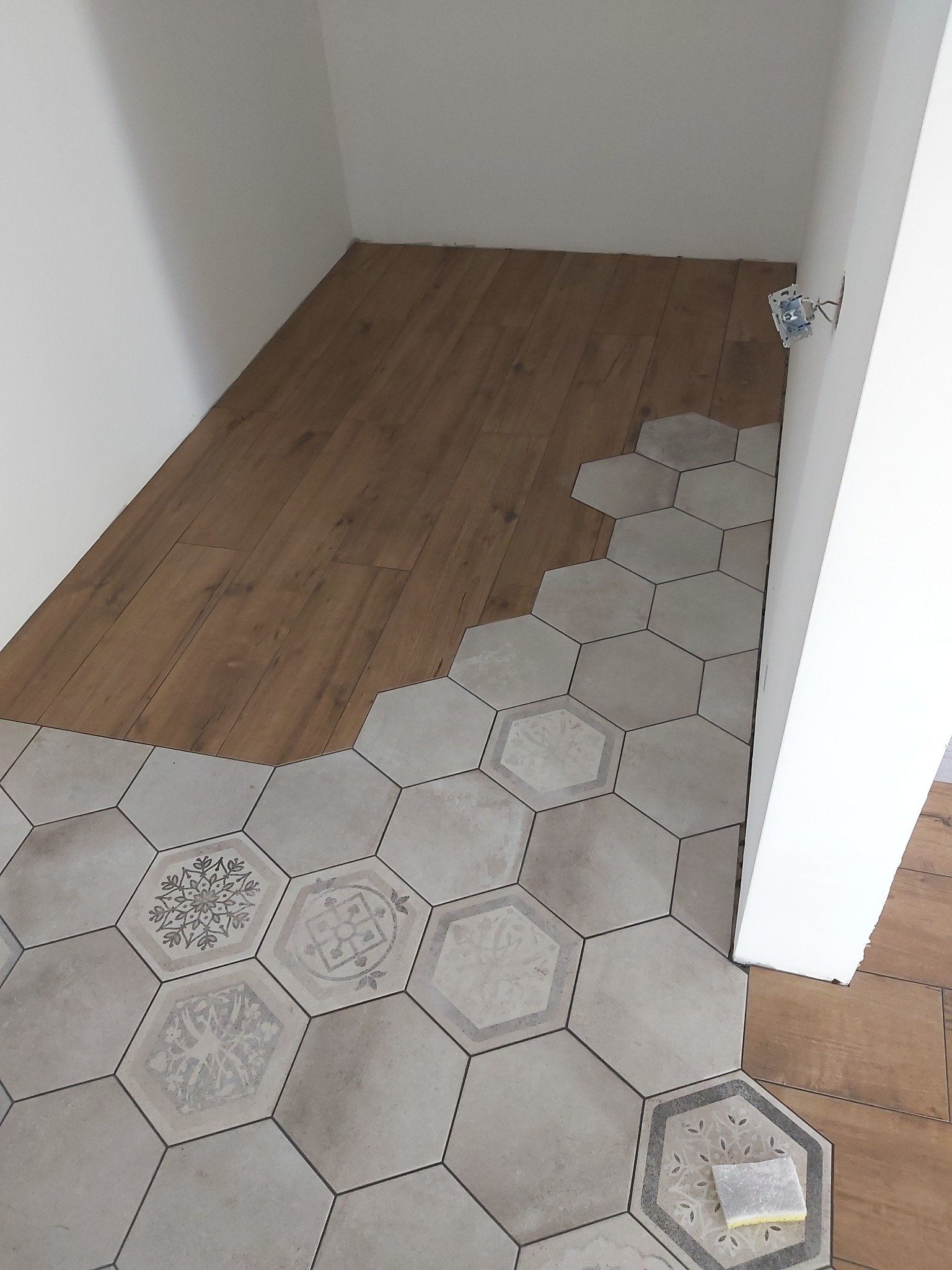 Układanie heksagonalnych płytek podłogowych z wzorami, częściowo widoczna podłoga drewnopodobna, biała ściana z wystającą puszką elektryczną.