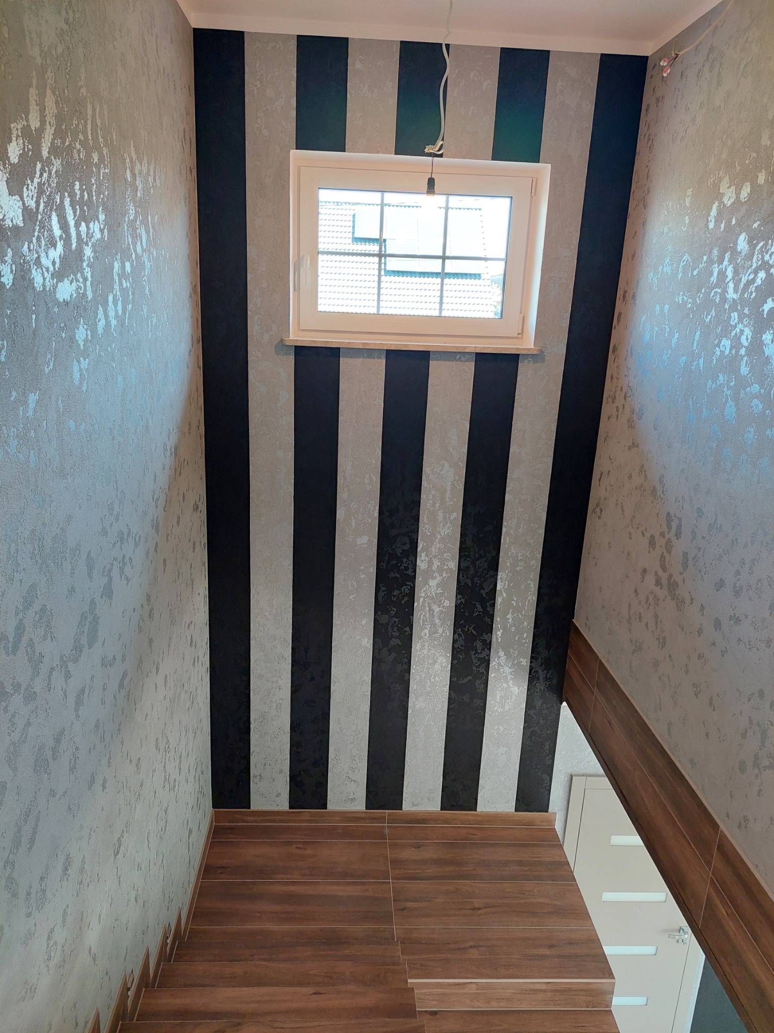 Klatka schodowa po renowacji: ściany z efektem metalicznego połysku i czarno-srebrne pasy, drewnopodobne schody. Okno doświetla przestrzeń.