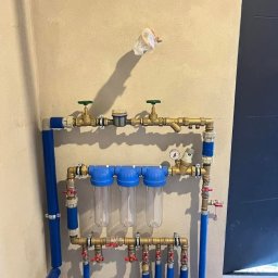 ABC Instal - Niebieskie rury i filtry wody zamontowane na ścianie, z zaworami i manometrem. Instalacja hydrauliczna w surowym wnętrzu, widoczne łączenia i elementy mocujące.