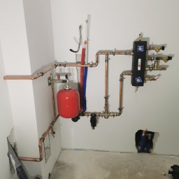 ABC Instal - Miedziana instalacja CO z czerwonym zbiornikiem wyrównawczym na białej ścianie w surowym wnętrzu. Widoczne liczne zawory i przyłącza. Elementy instalacji w trakcie montażu.