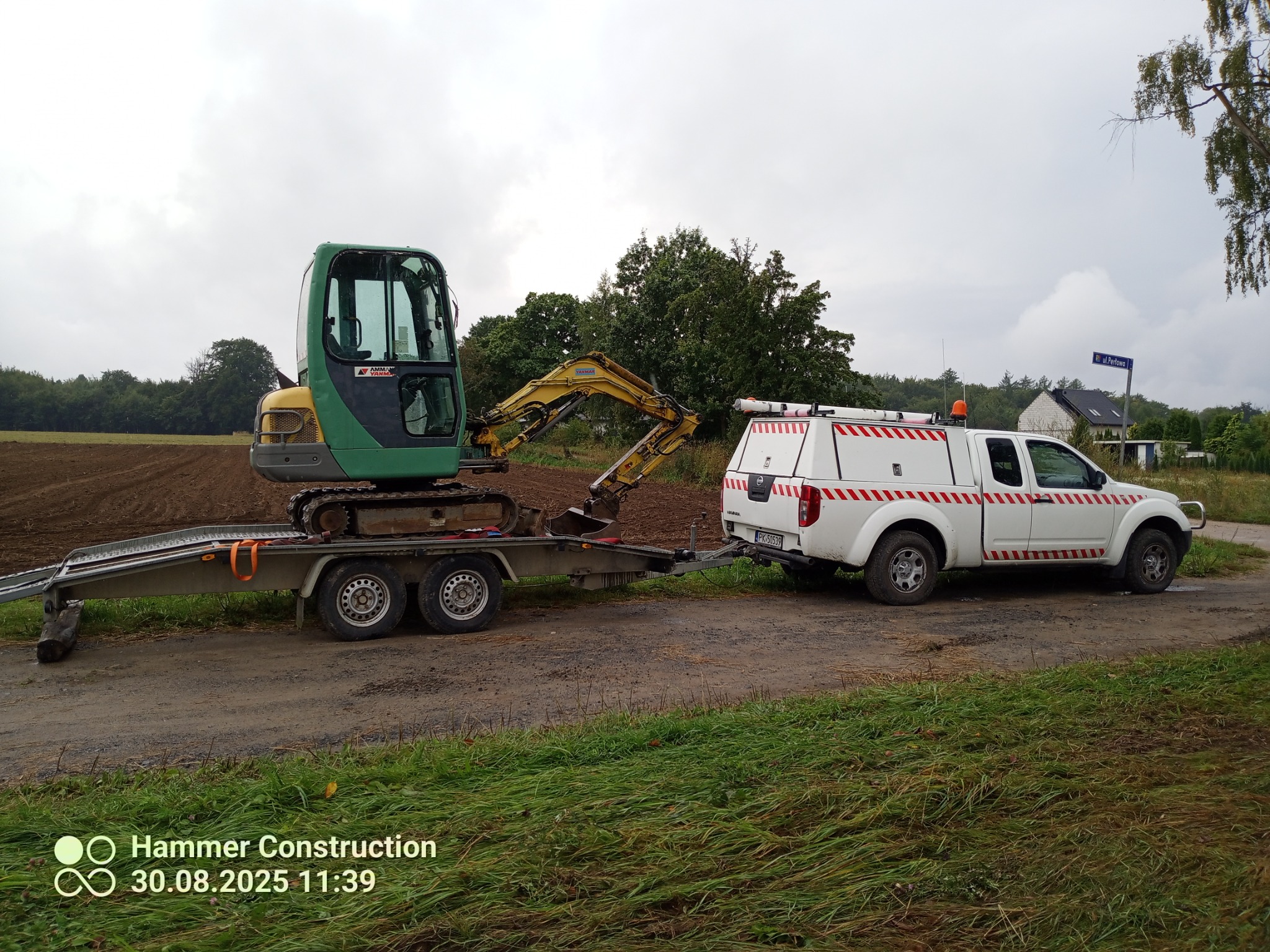 Transport mini koparki Yanmar na przyczepie, ciągniętej przez białego pickupa Nissan Navara z zabudową. Tło: pole, drzewa, pochmurne niebo.