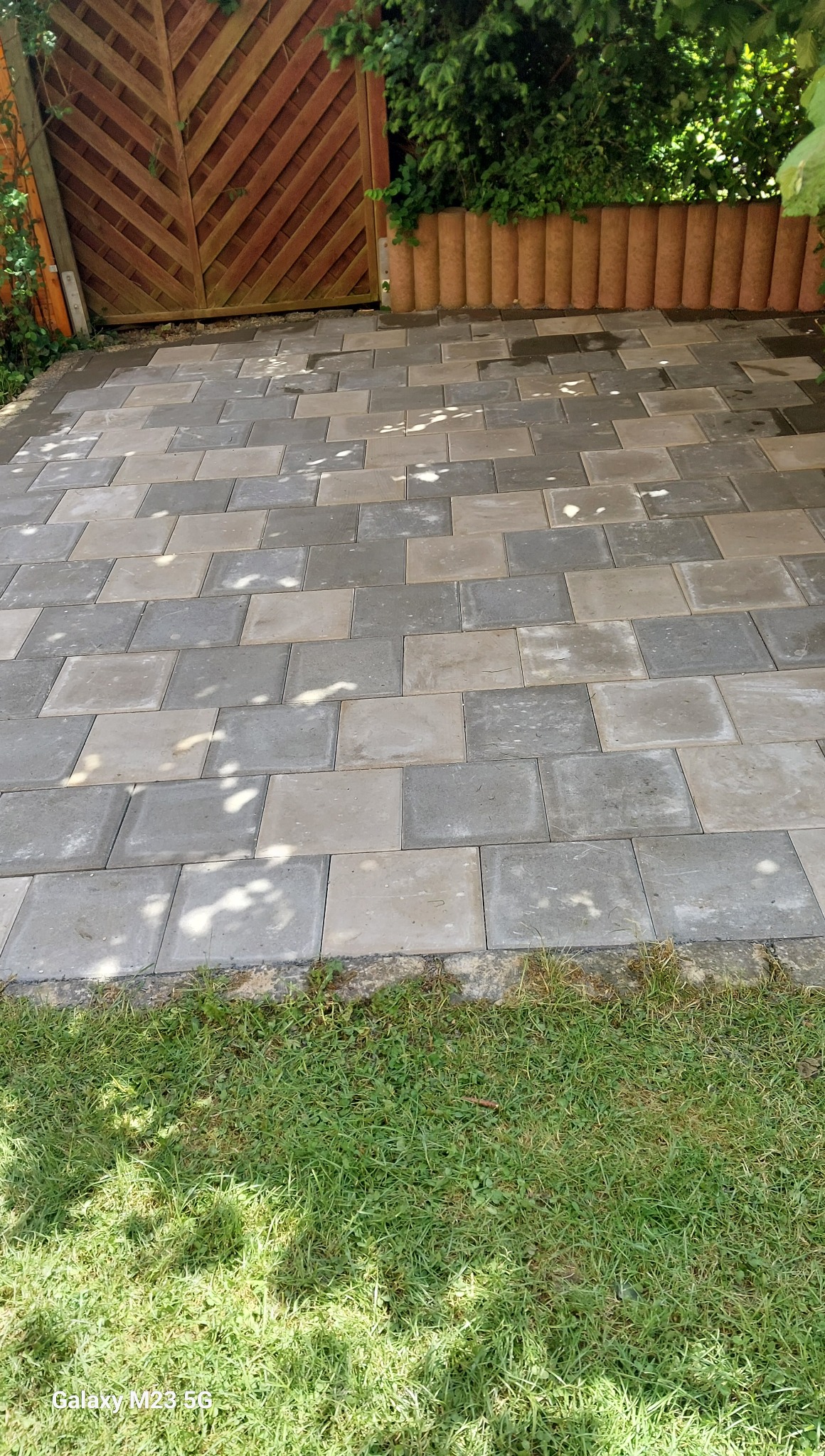 Ułożona szara kostka brukowa na podwórku, widok z góry, z drewnianą bramą w tle i zielenią po bokach. Cień drzew na kostce.