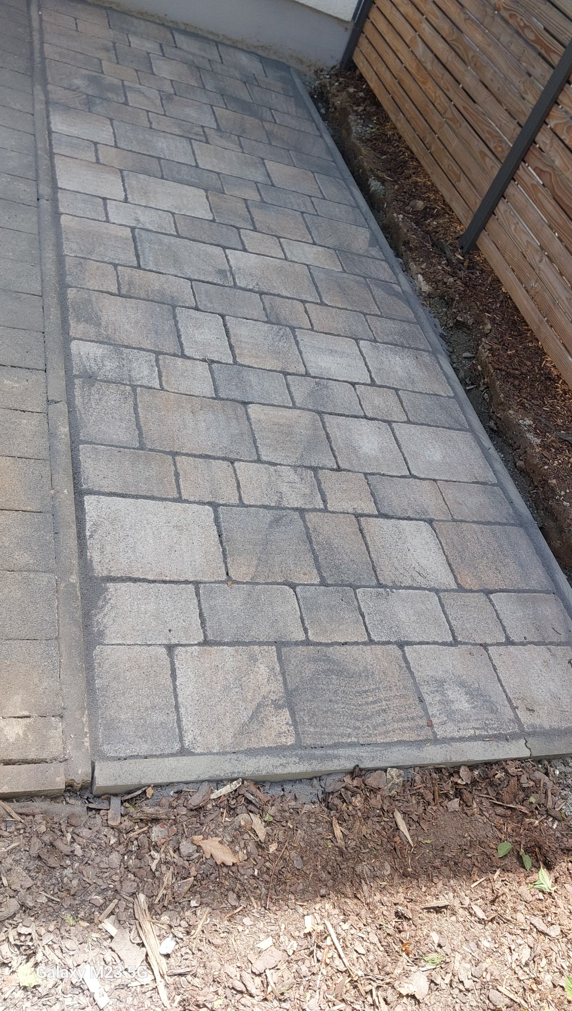 Ułożona kostka brukowa w szarym odcieniu, widok z góry. Krawędź kostki graniczy z drewnianym płotem i rabatą z korą. Widoczny fragment budynku w tle.