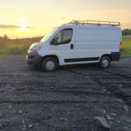 Biały bus z relingami dachowymi na żwirowym podłożu, w tle łąka i zachodzące słońce. Pojazd zaparkowany bokiem, oświetlony ciepłym światłem.