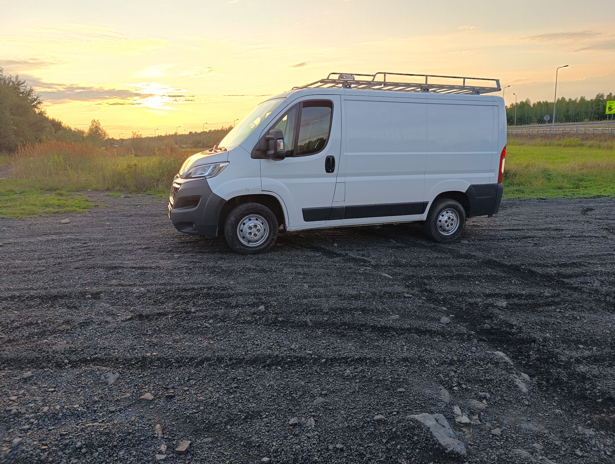Biały bus z relingami dachowymi na żwirowym podłożu, w tle łąka i zachodzące słońce. Pojazd zaparkowany bokiem, oświetlony ciepłym światłem.