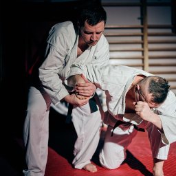 Tomasz Misiak - Trening sportów walki: mężczyzna w białym kimonie wykonuje dźwignię na stawie łokciowym przeciwnika na czerwonej macie w sali z drabinkami.