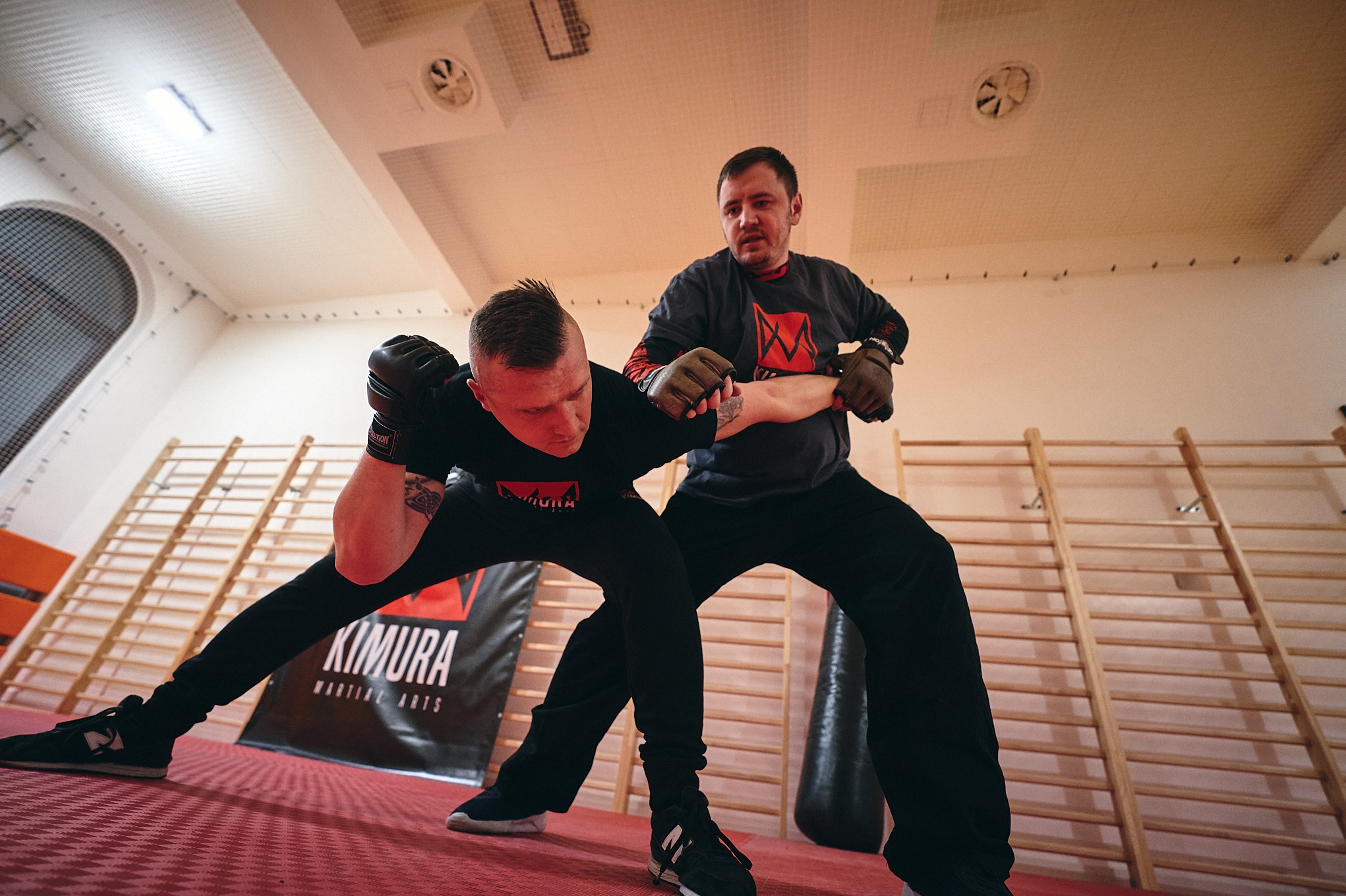 Trening sportów walki w sali: mężczyzna w czarnym stroju ćwiczy chwyt pod okiem trenera na tle drabinek i baneru Kimura Martial Arts.