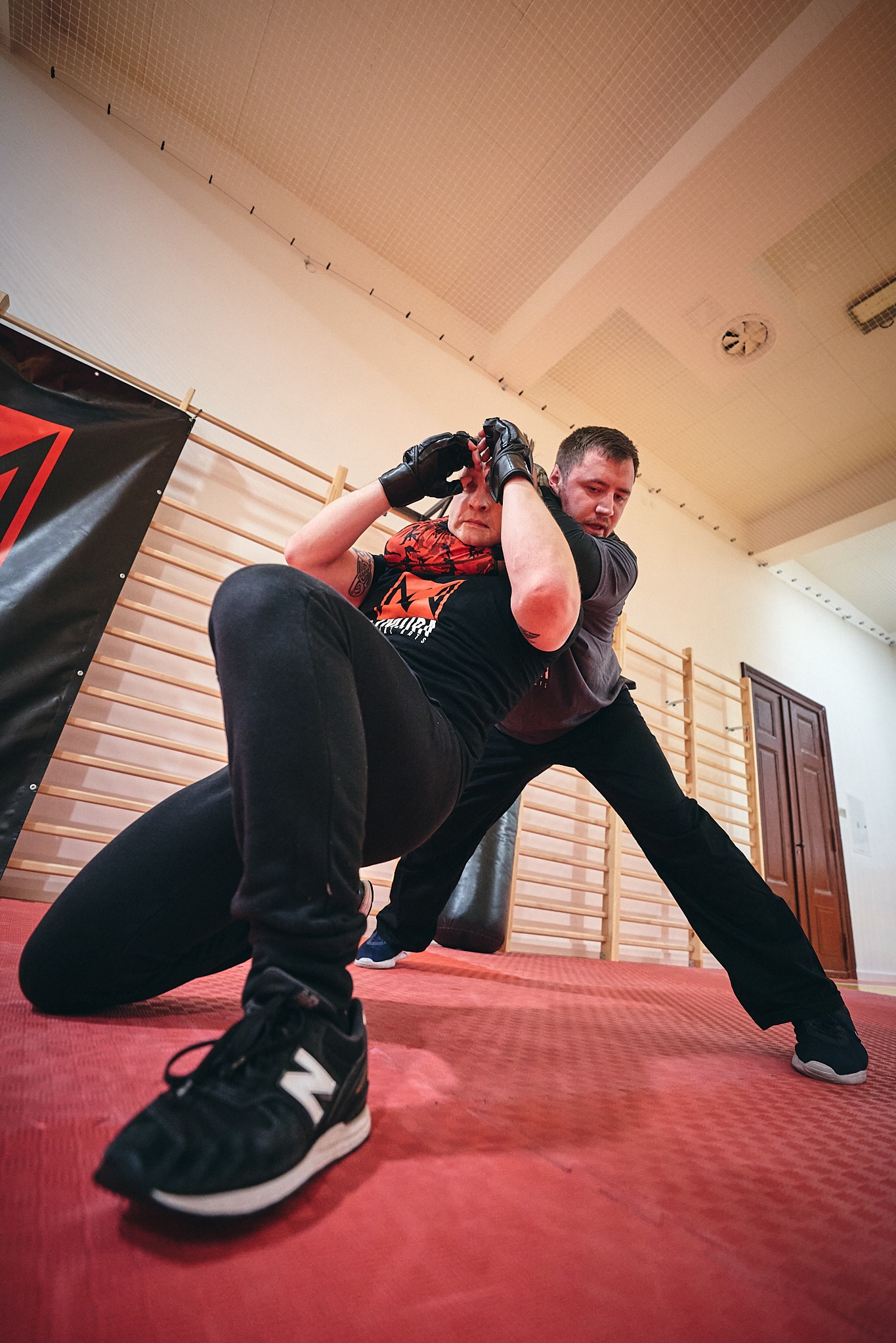 Trening samoobrony: mężczyzna uczy kobietę chwytu obronnego w sali sportowej z drabinkami i workiem bokserskim. Kobieta w rękawicach, ujęcie z dołu.