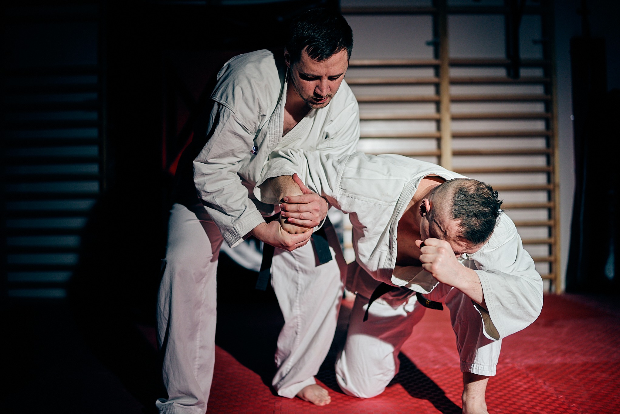 Trening sportów walki: mężczyzna w białym kimonie wykonuje dźwignię na stawie łokciowym przeciwnika na czerwonej macie w sali z drabinkami.
