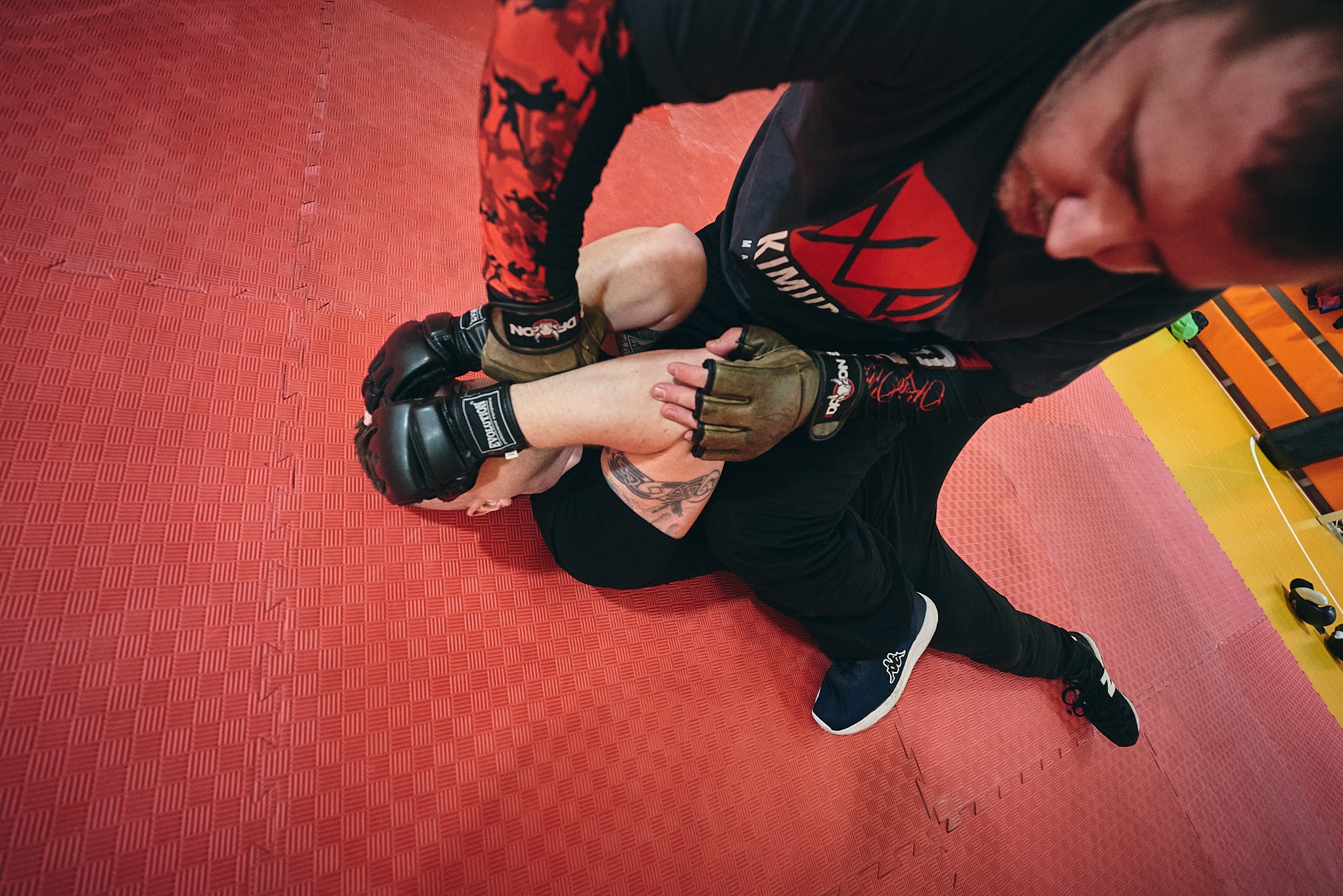 Zawodnik MMA w czarnej odzieży, w trakcie duszenia przeciwnika na czerwonej macie. Widoczne tatuaże i rękawice do MMA. Kąt ujęcia z góry.