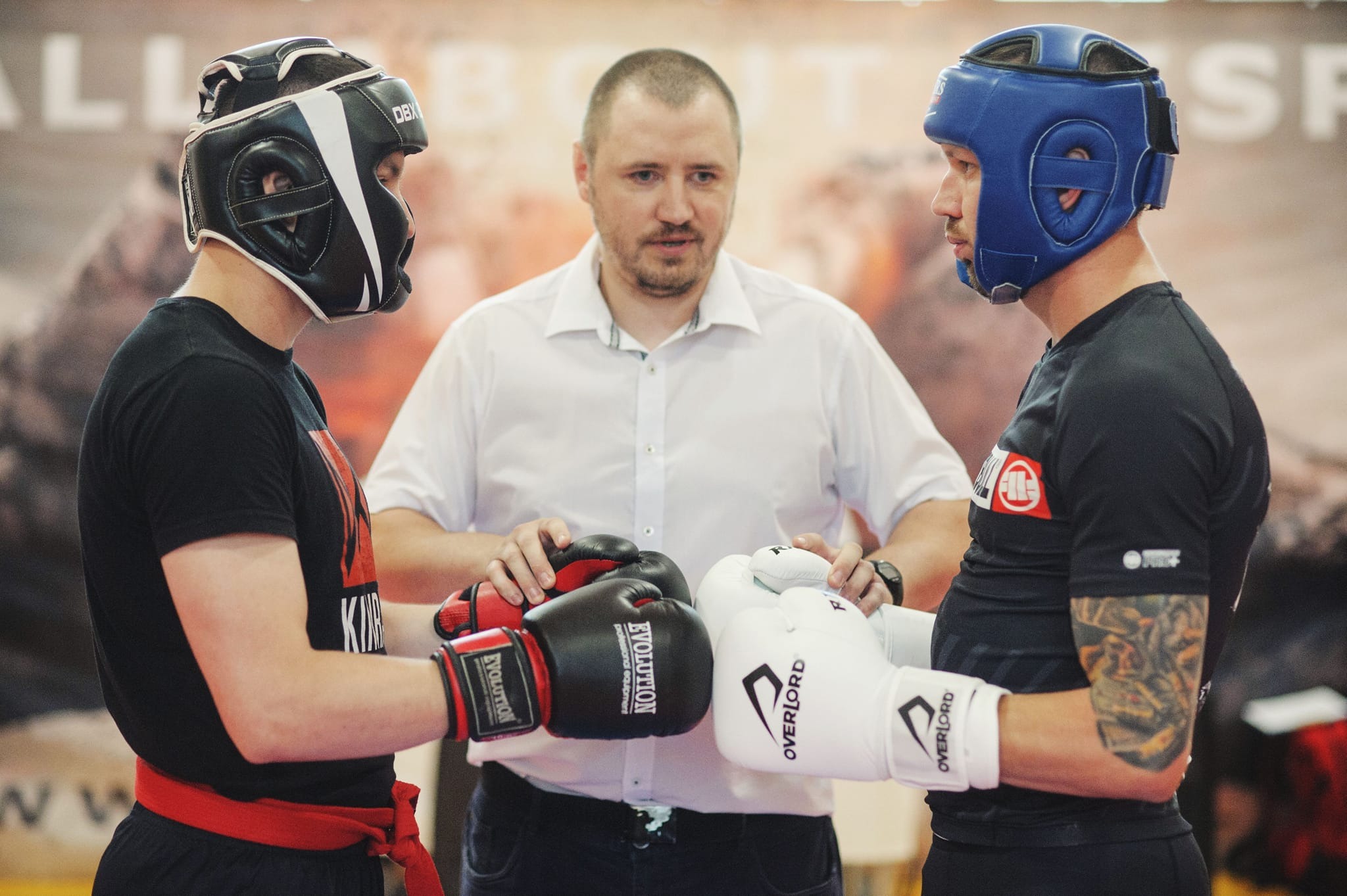 Dwóch zawodników w kaskach i rękawicach bokserskich stoi naprzeciwko siebie, nadzorowanych przez trenera. Widoczne logo na rękawicach i strojach.