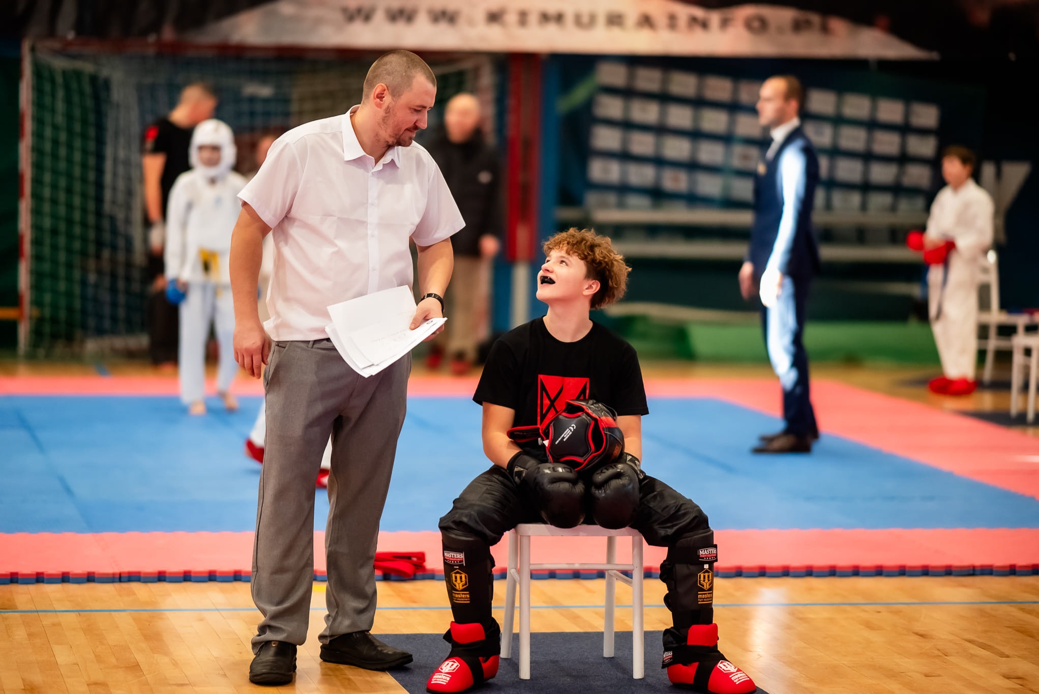 Trener personalny z kartką rozmawia z młodym zawodnikiem sportów walki w pełnym stroju ochronnym, siedzącym na krześle. W tle inni zawodnicy i trenerzy.