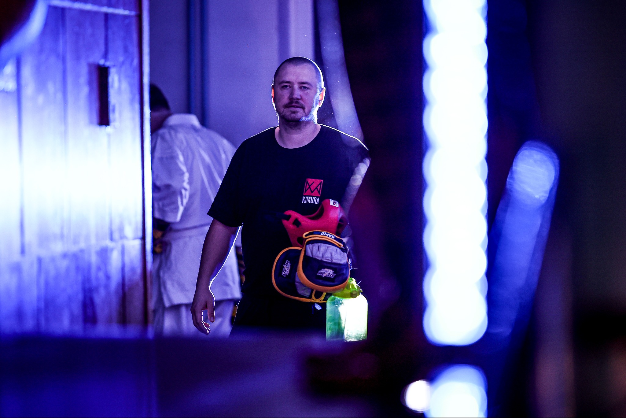 Mężczyzna w czarnej koszulce z logo Kimura, trzymający rękawice bokserskie i ochraniacz, na tle sali treningowej z oświetleniem neonowym.