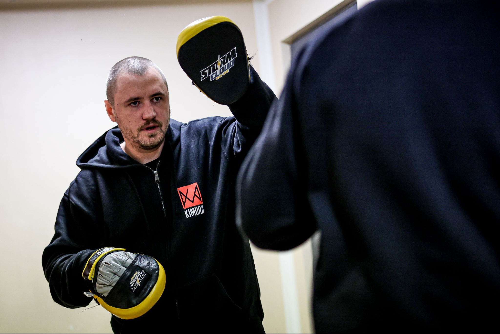 Mężczyzna w czarnej bluzie z logo Kimura, z ochraniaczami na dłoniach, trenuje sporty walki z niewyraźną postacią w tle. Dynamiczne ujęcie treningu.