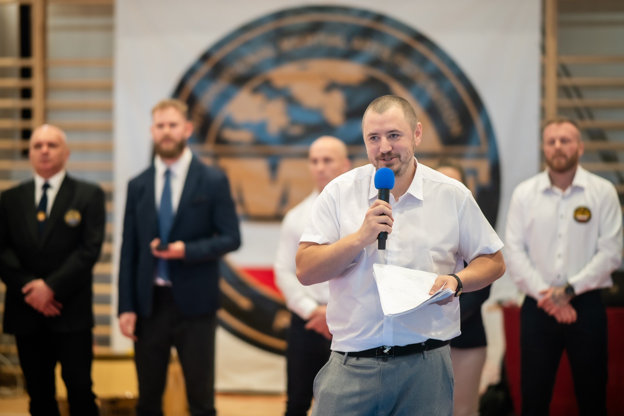 Prelegent z mikrofonem na tle logo organizacji sportów walki, otoczony przez osoby w strojach formalnych podczas wydarzenia.