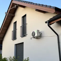 Zewnętrzna jednostka klimatyzacji KAISAI na elewacji domu z oknami balkonowymi i brązową dach&oacute;wką. Widoczna antena na dachu i rynna. Biała ściana, błękitne niebo.