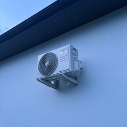 Dawid Tomaszewski - Zewnętrzna jednostka klimatyzacji Kaisai zamontowana na białej ścianie budynku pod ciemnym okapem. Widoczne mocowania i przewody. Delikatne oświetlenie.