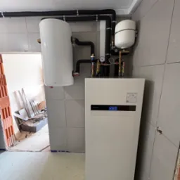 Biała pompa ciepła Panasonic Aquarea Smart Cloud z zbiornikiem buforowym i rurami w izolacji, zamontowana na szarej ścianie z płytek, widok na remontowane pomieszczenie.